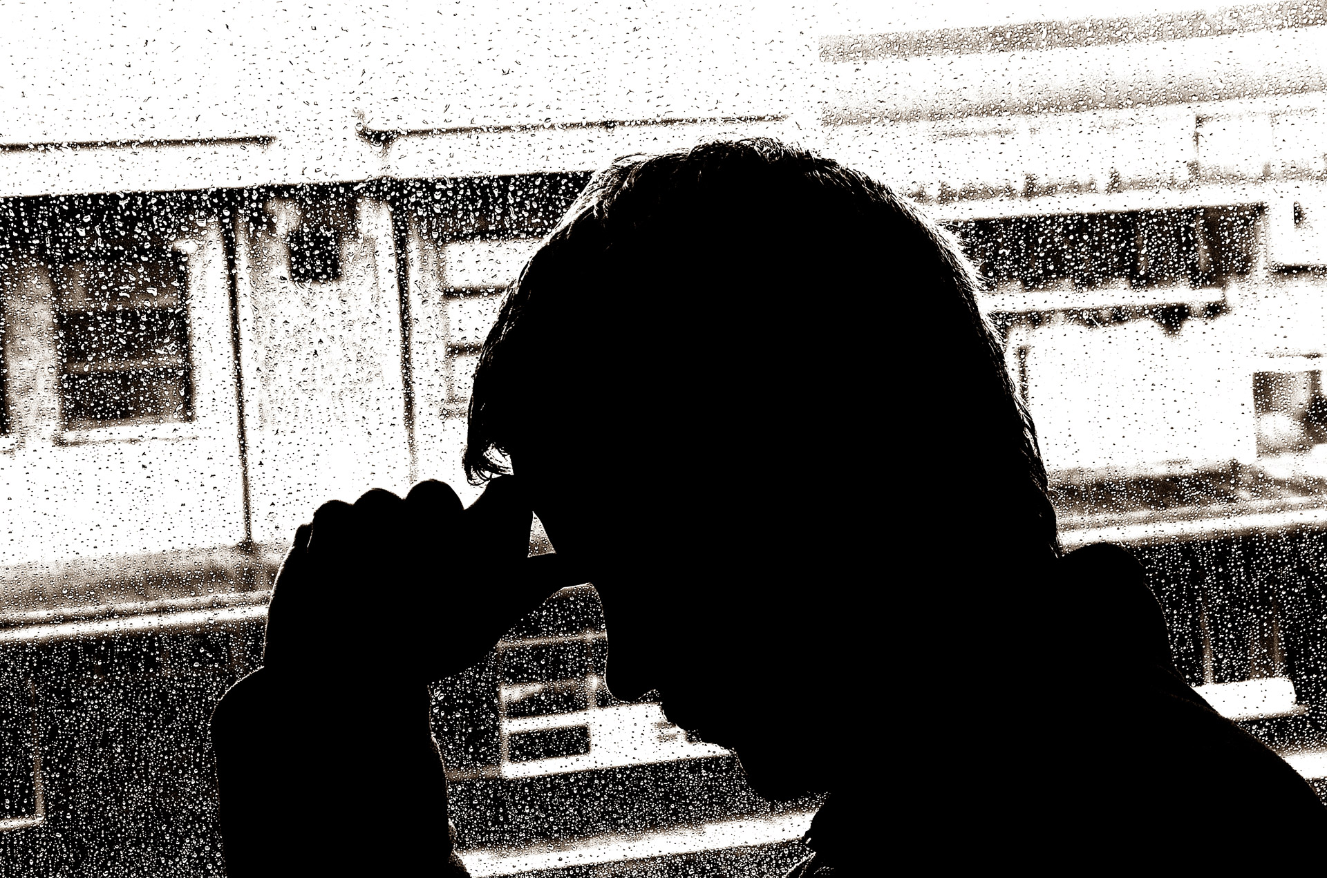 The silhouette of a man looking out a window with his hand across his forehead, as if having depression.